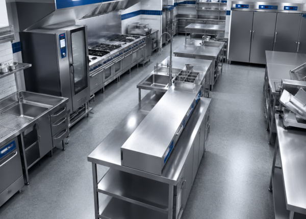 Industrial commercial kitchen with stainless steel worktables, ovens, and refrigeration along white-and-blue tiled walls under brightPendant lights.